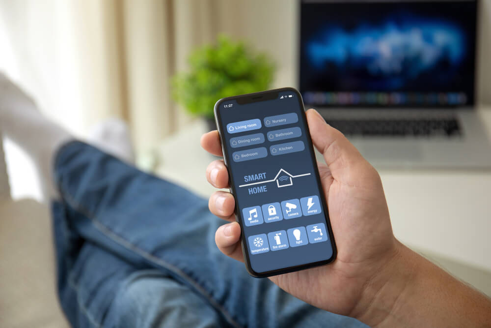 Man on Sofa Holding Phone With App Smart Home on Screen in Room House