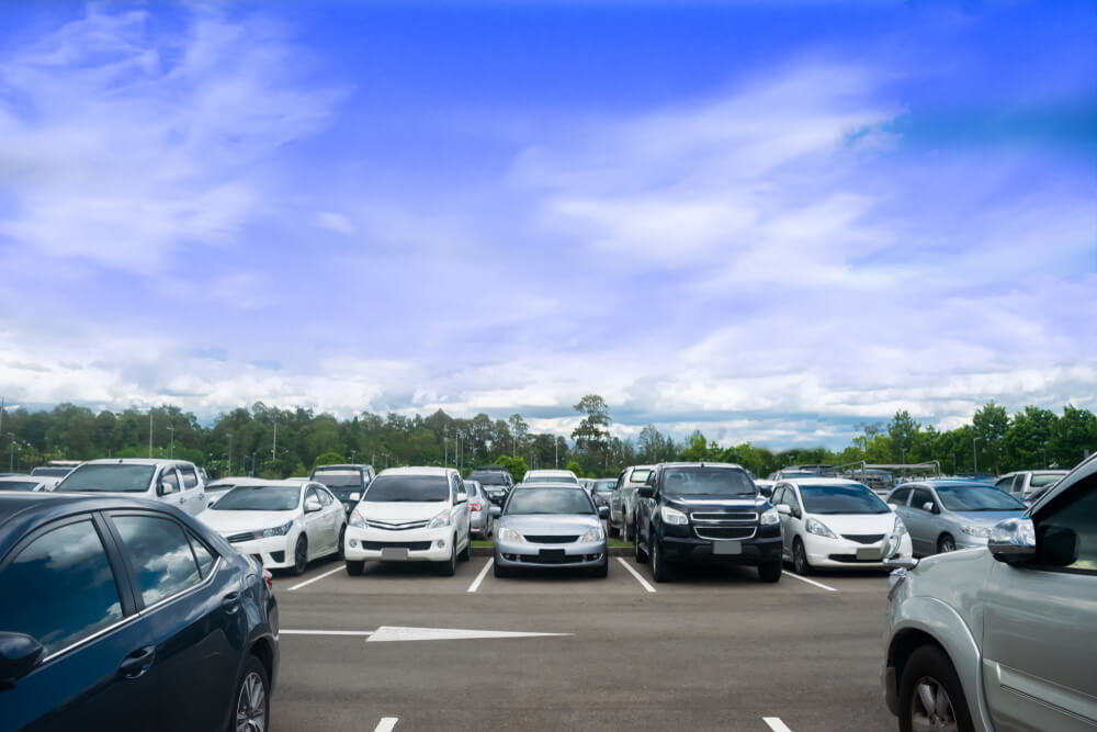 Car Parking in Large Asphalt Parking Lot With Trees, White Cloud and Blue Sky Background